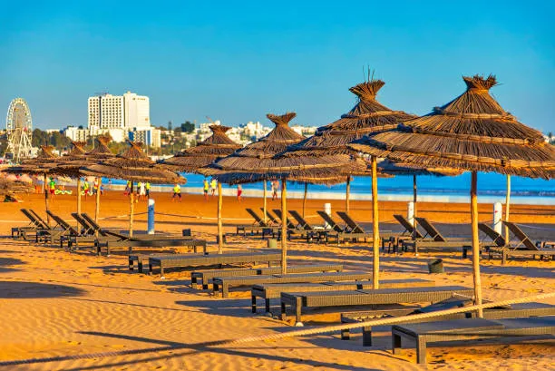 Straw beach umbrellas and sun loungers on the golden sand of Agadir Beach along the Atlantic Ocean in Morocco, with the city skyline and seaside promenade in the background under a clear blue sky.